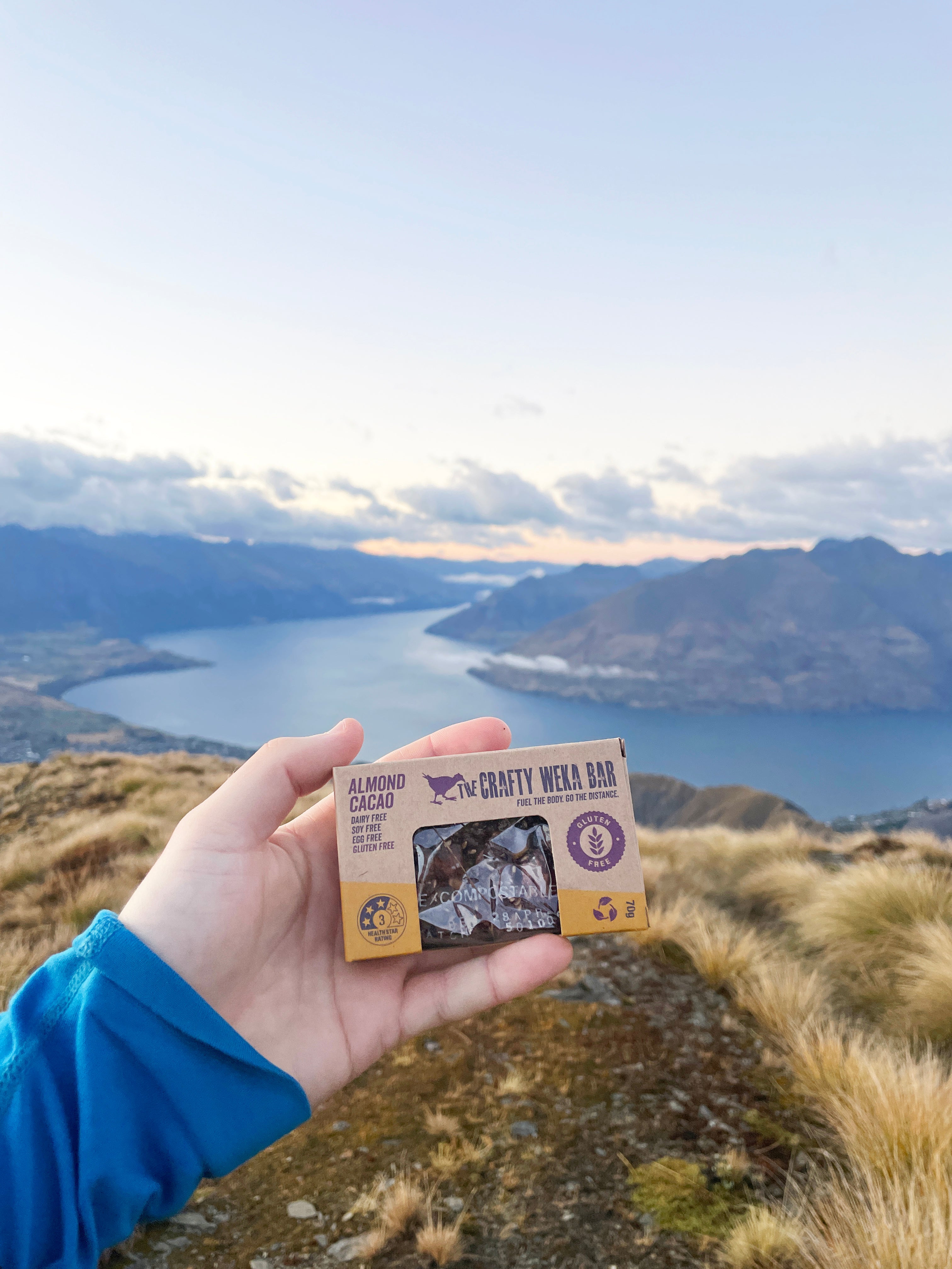 Hand holding a 'The Crafty Bar' snack bar with a scenic mountain and lake background